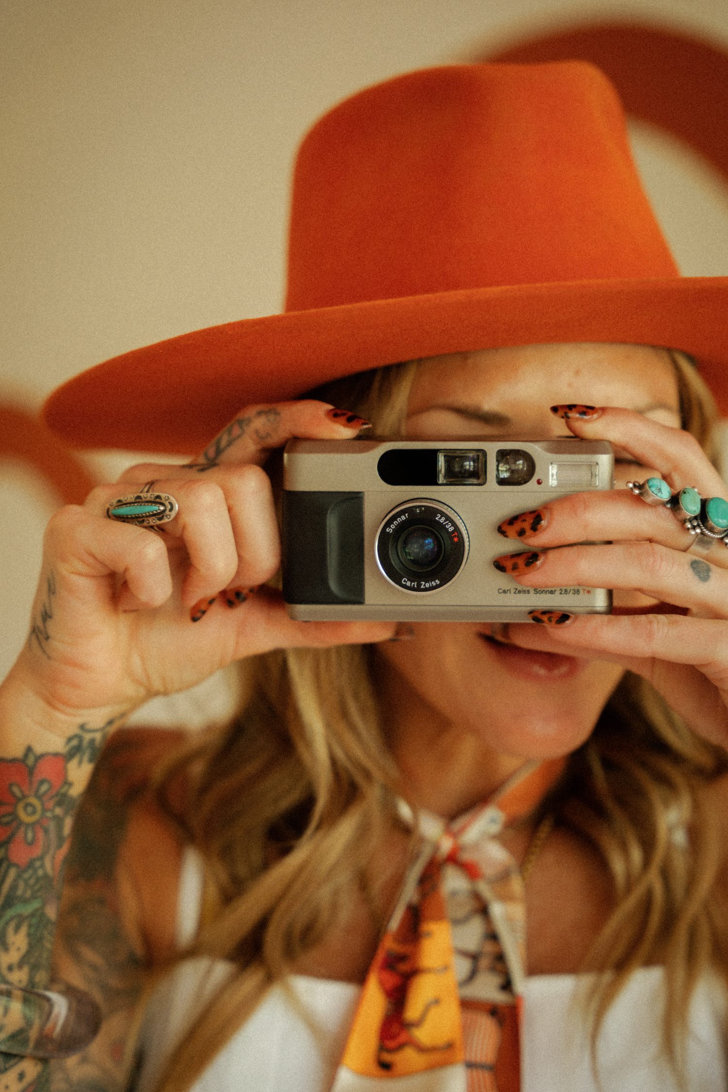 Leopard-print nails and turquoise rings holding a vintage Carl Zeiss film camera under a terracotta hat